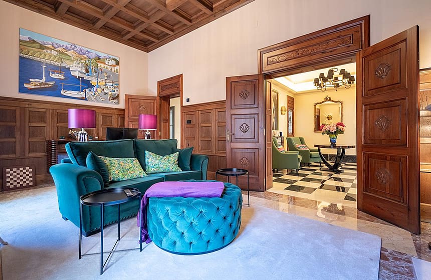 A formal living room with dark wood wall paneling, a coffered ceiling, a velvet teal sofa, and an open doorway leading to a tiled room.