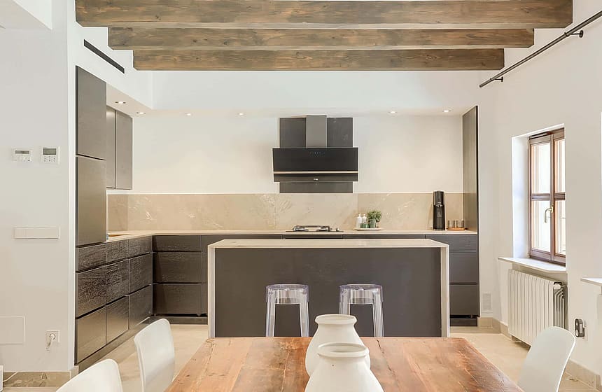 Modern kitchen with dark cabinetry, marble backsplash, central island, and wood dining table under exposed ceiling beams.