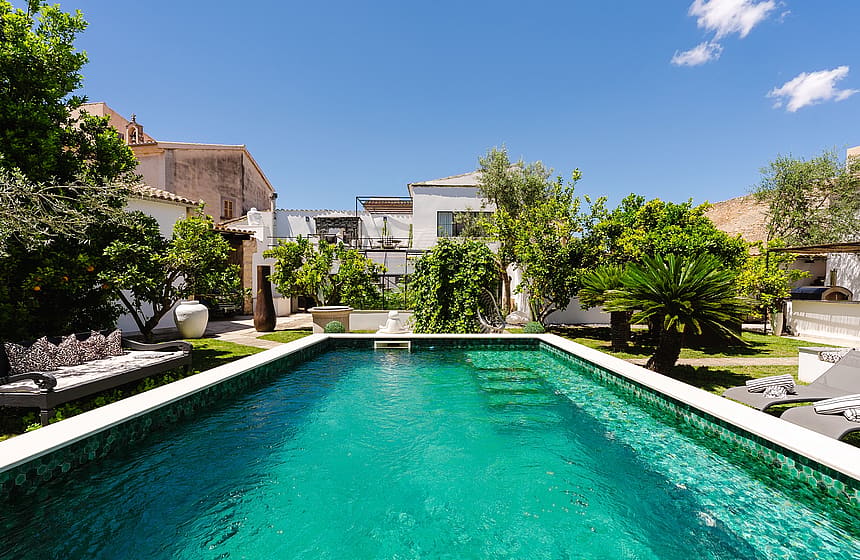 A rectangular swimming pool with turquoise mosaic tiles set within a lush, walled garden in Sencelles, featuring mature trees.