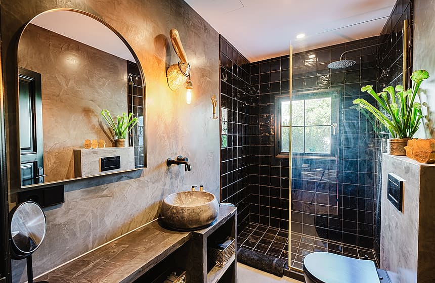 Contemporary bathroom with grey concrete walls, a dark stone washbasin, arched mirror, and a black tiled walk-in shower.