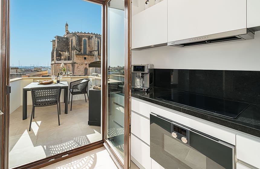 Modern white kitchen with black countertops opening to a sunny terrace with historic church views in Palma Old Town.