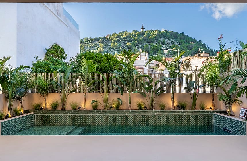 A rectangular pool with green chevron tiling and tropical plants, set against a backdrop of the Na Burguesa hills.