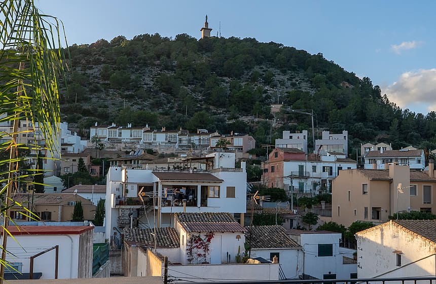 An elevated view across white Mediterranean rooftops toward a green hillside topped with a stone monument.