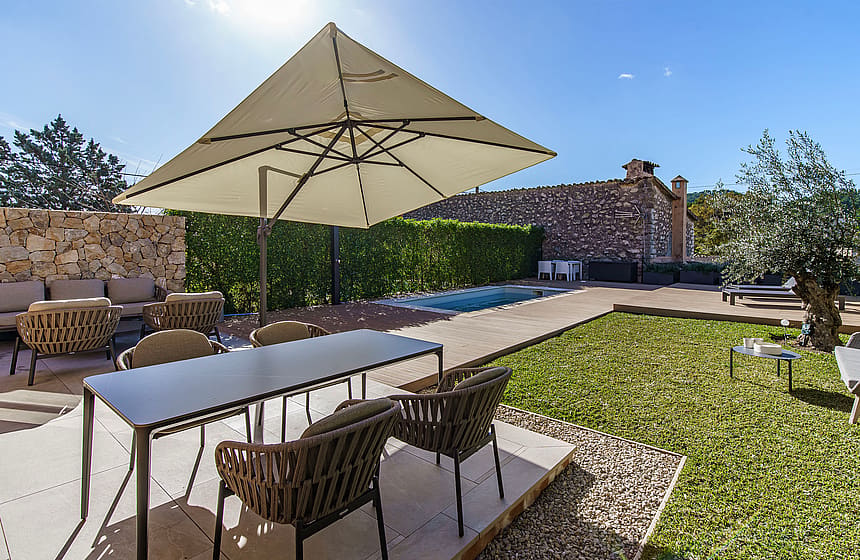 An outdoor dining terrace in Es Capdella featuring a rectangular table with woven chairs, a large beige parasol, and a small pool set against a natural stone wall.