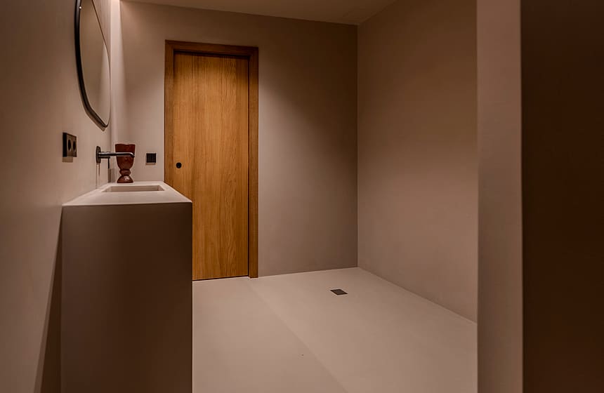 A minimalist bathroom featuring a white monolith vanity, a circular mirror, and a natural wood door under warm lighting.