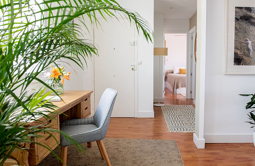 A home office area with a light wood desk, grey upholstered chair, and potted plant in the foreground, leading to a bedroom.
