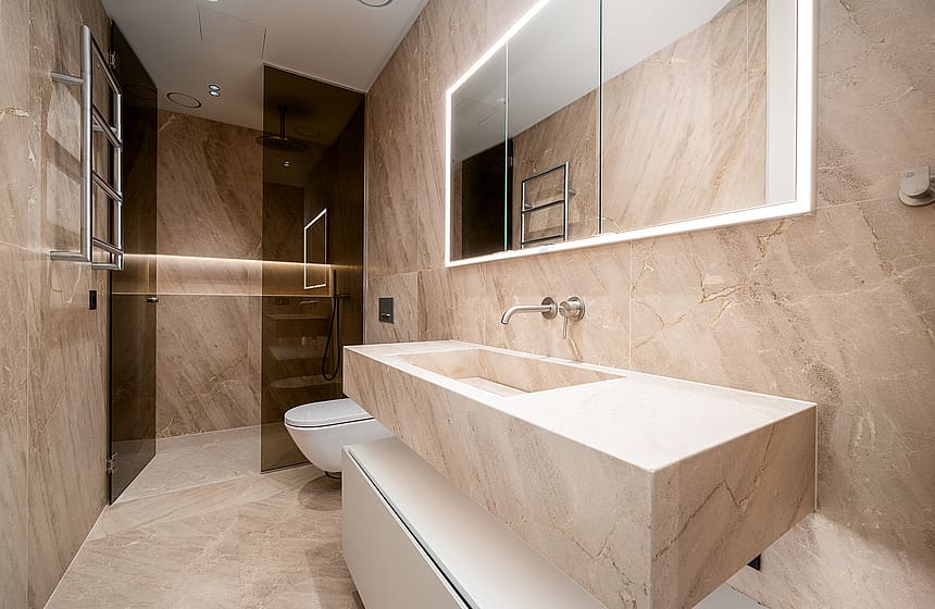 Modern bathroom featuring beige stone wall cladding, a floating stone vanity sink, a wall-mounted toilet, and a glass-enclosed shower area with integrated LED mirror lighting.