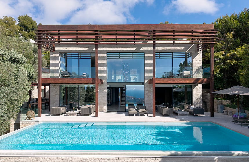 Exterior of a contemporary villa featuring an infinity pool, natural stone facade, floor-to-ceiling glass, and timber pergola.