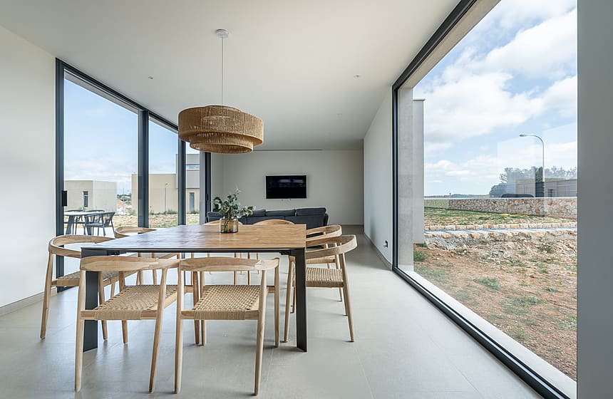 Dining room with a wooden table, woven chairs, and large glass windows overlooking a dry landscape.