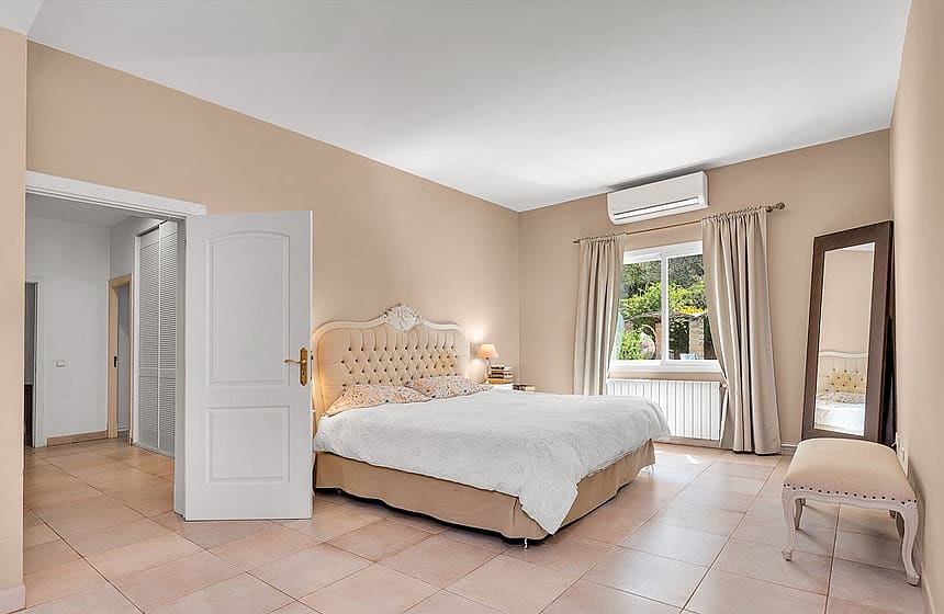 A spacious bedroom featuring a tufted upholstered bed, light beige walls, tiled floors, and a floor mirror.