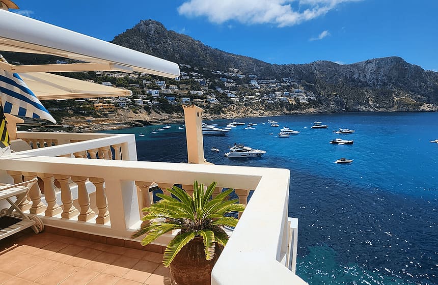 A wide-angle shot of a terrace with white balustrades overlooking the blue waters of Cala Llamp, featuring mountains and anchored boats.