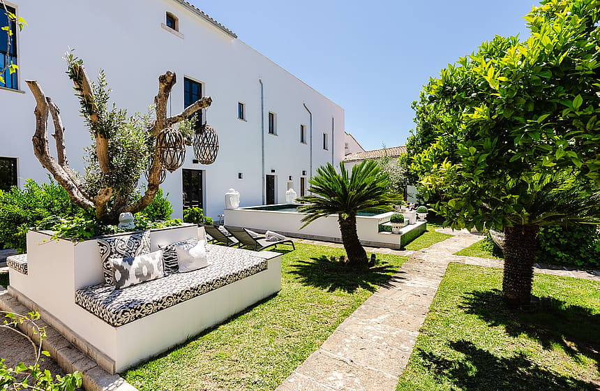 A white townhouse exterior with a landscaped garden featuring a built-in stone bench, patterned cushions, and a palm tree.