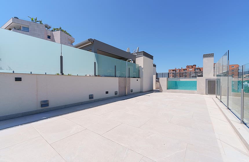 Terraza espaciosa en azotea con baldosas de piedra, delimitada por paneles de cristal bajo un cielo azul.