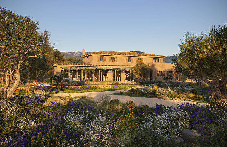 A two-story stone finca set behind a Mediterranean garden with purple wildflowers and olive trees under a clear sky.