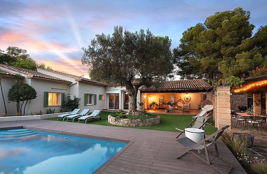 Outdoor swimming pool with wood decking, lounge chairs, and a central olive tree in a villa garden during dusk.