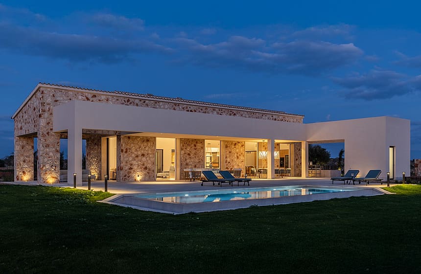 Wide shot of a stone and white plaster finca at dusk, featuring a lit swimming pool and patio furniture on the terrace.