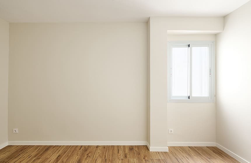 Empty bedroom with light beige walls, wooden floor, and a single white-framed window set into a niche.