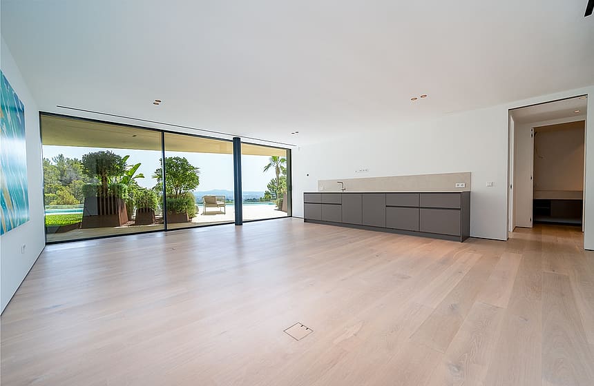 An expansive, unfurnished living room with wide plank light oak flooring, white walls, and a low grey modular cabinet. Floor-to-ceiling glass sliding doors open to a sunlit terrace with greenery and distant sea views.