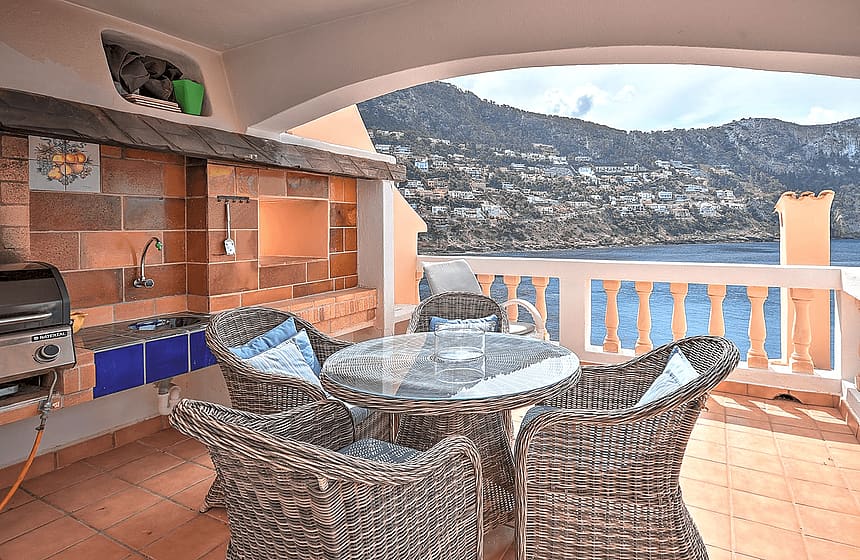 Covered terrace featuring an outdoor brick barbecue and kitchen area with dining table and wicker chairs overlooking the sea.