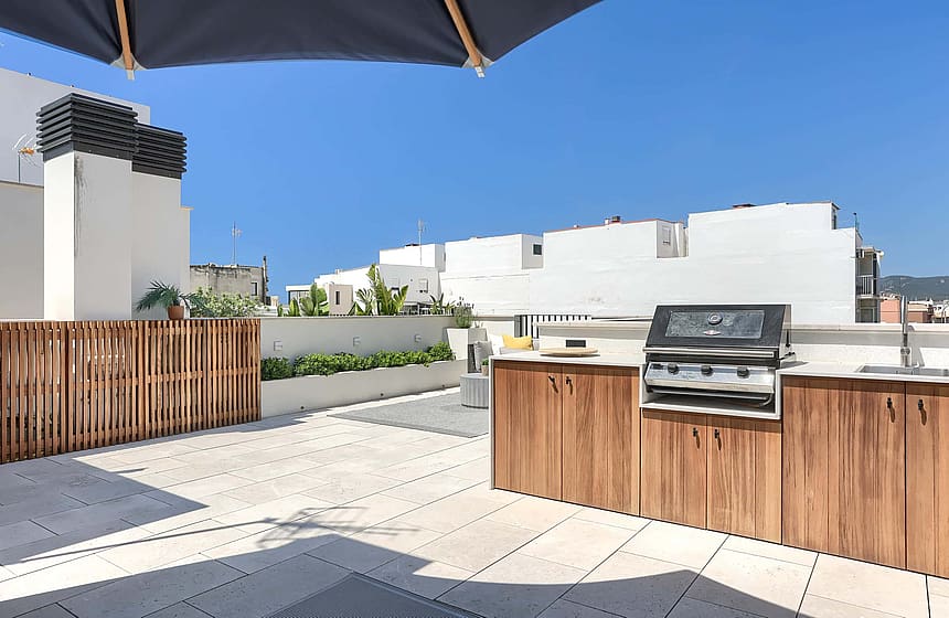 Rooftop terrace featuring a wooden outdoor kitchen cabinet with a built-in stainless steel grill, stone tile flooring, and slatted wood privacy fencing under a clear blue sky.