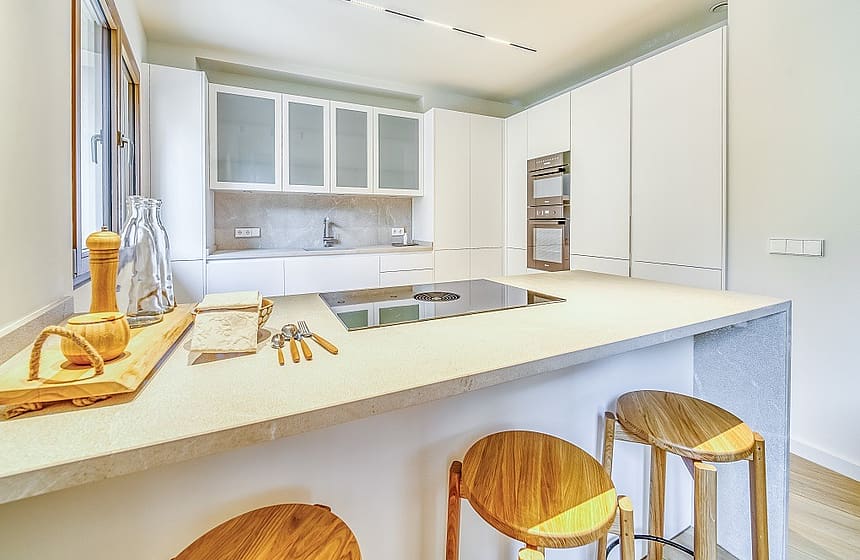 Modern kitchen with white cabinetry, integrated appliances, and a light stone island with wooden bar stools in the foreground.