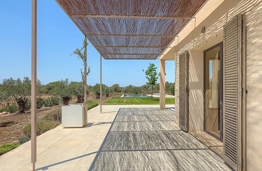 A wide outdoor terrace featuring a woven reed pergola casting dappled shadows on grey stone flooring, next to the finca wall.