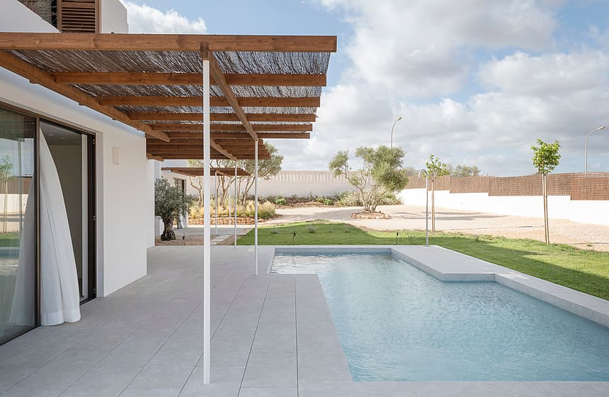 Modern pool terrace featuring white stone tiling and a timber pergola with reed shading next to a garden lawn.