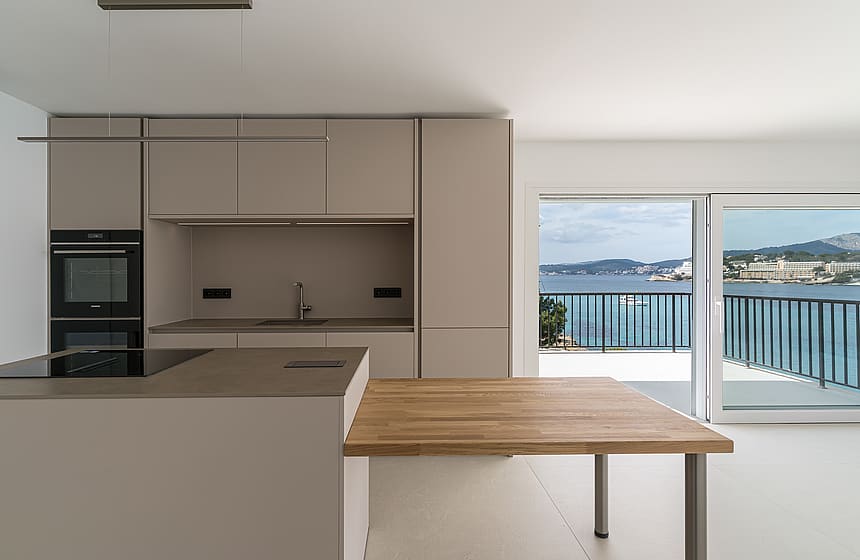 Minimalist kitchen with taupe cabinetry and an oak dining surface opening onto a sea-view terrace.
