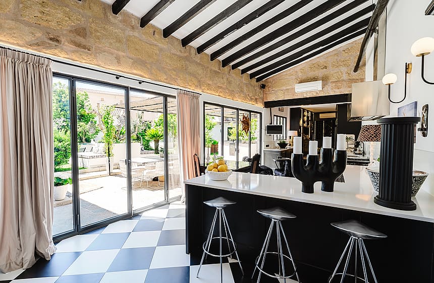 Modern kitchen with black cabinetry, white countertops, checkered floors, and exposed stone walls under a high beamed ceiling.