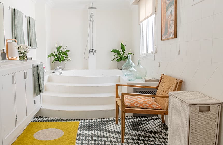 A bright bathroom with white walls, a stepped white bathtub, a woven wood chair, and patterned floor tiles.