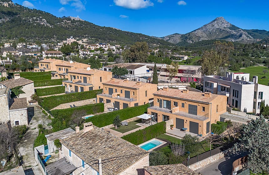 Aerial drone view showing a row of terraced townhouses with ochre facades, landscaped gardens, and a pool in Es Capdella.