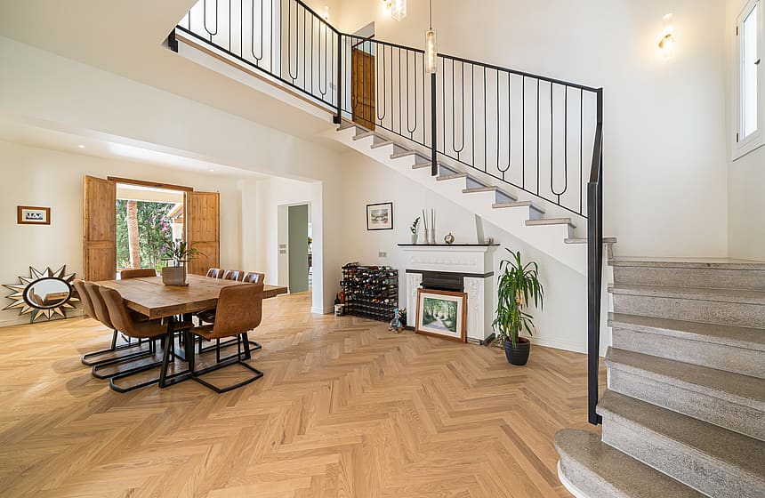 Sunlit living space with oak chevron floors, a black iron staircase, and a dining table near open wooden shutters.