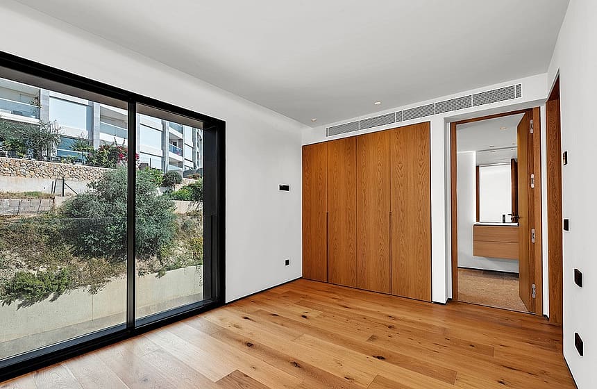 A minimalist bedroom featuring light oak flooring, large integrated oak wardrobes, and a sliding glass door looking out to greenery.