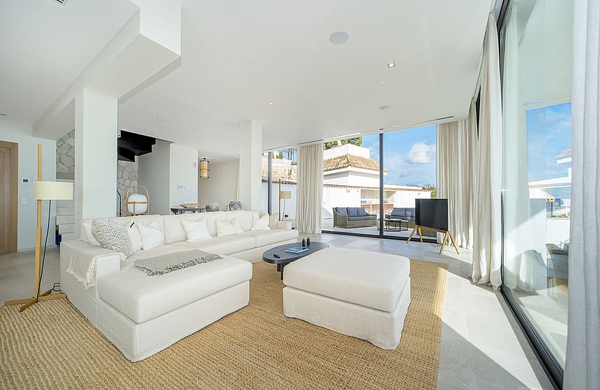 Bright living room with white L-shaped sofa, matching ottoman, woven area rug, and floor-to-ceiling glass sliding doors.