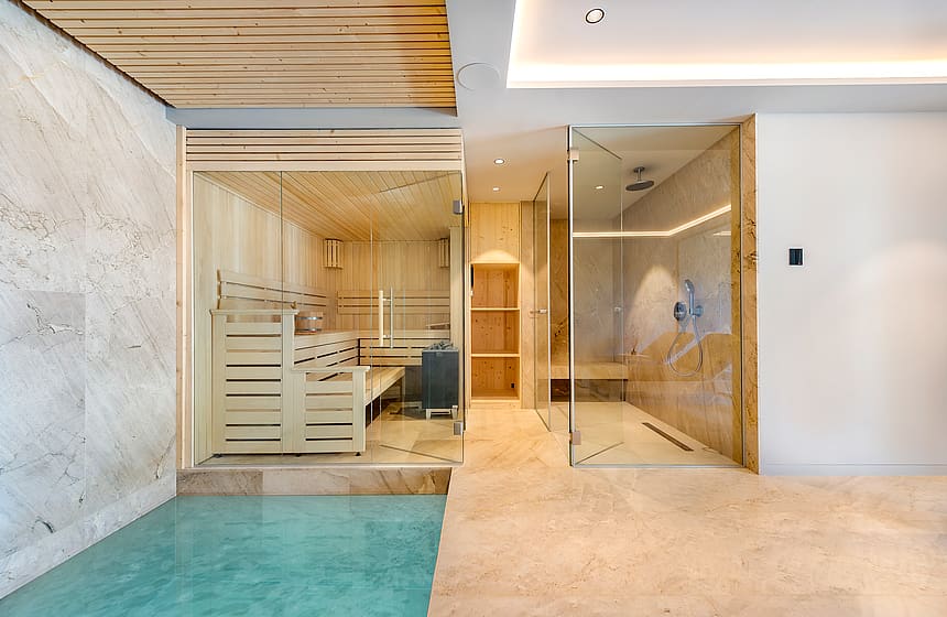 Modern home spa featuring a glass-enclosed cedar sauna beside a walk-in rainfall shower with stone flooring.