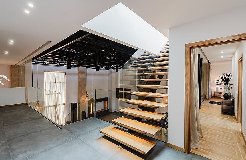 A modern floating oak staircase with glass side panels, illuminated by ceiling spotlights and a skylight.