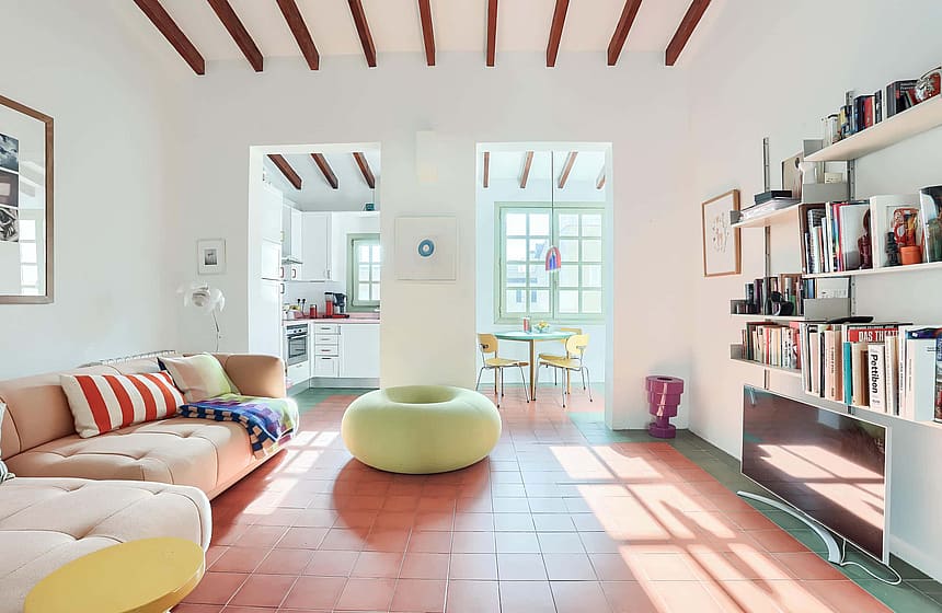 A bright living area featuring terracotta floor tiles, exposed wooden ceiling beams, a sofa with a striped cushion, and a round lime green ottoman, looking through to a dining area.