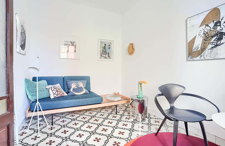 A bright living space featuring a blue sofa, a dark designer chair, and traditional patterned floor tiles.