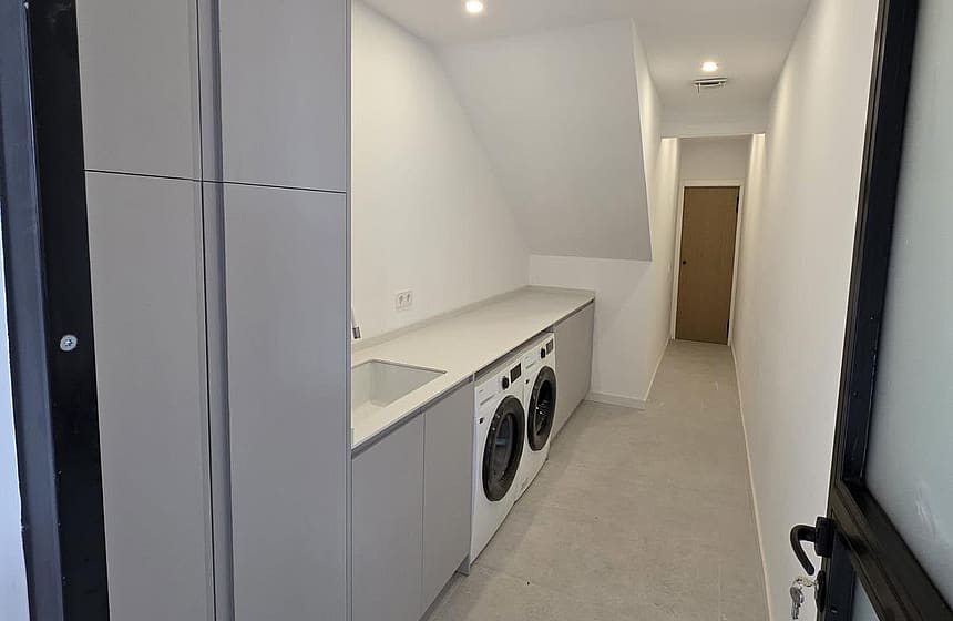 A narrow, minimalist laundry room with white cabinetry, integrated appliances, and light grey tile flooring.
