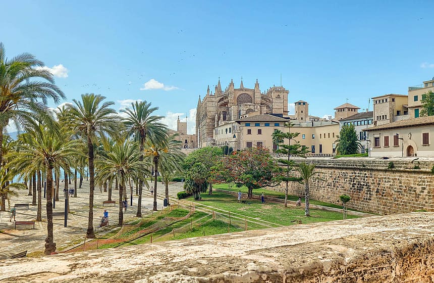 Panoramic view of a historic stone building and cathedral surrounded by palm trees and green gardens under a clear blue sky.