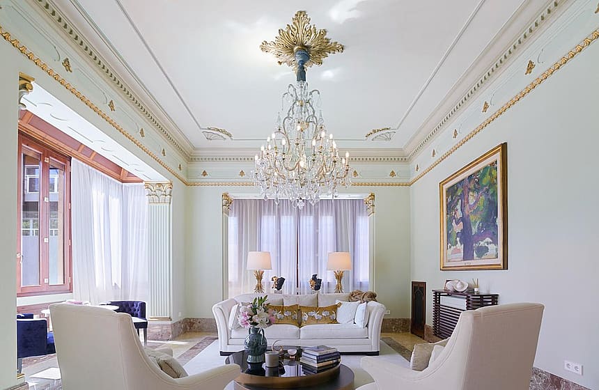 A formal living room with high ceilings, decorative gilded molding, and a crystal chandelier above a white sofa.