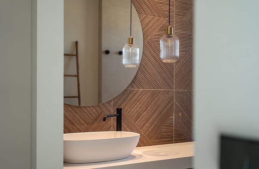 Close-up of a bathroom vanity featuring a white vessel sink, matte black faucet, and circular mirror against a textured brown tiled wall with glass pendant lights.
