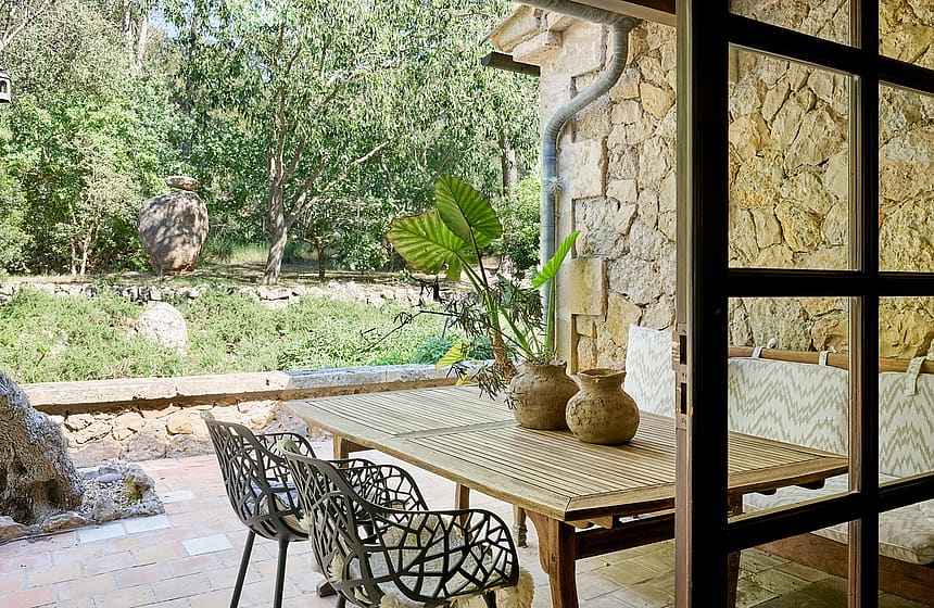 Outdoor dining set on a stone-paved terrace next to a rustic rubble stone wall, looking out to garden greenery.