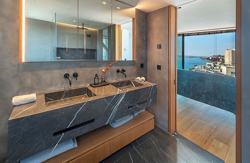 A modern bathroom featuring a grey marble double vanity with matte black faucets, a large mirror, and a view to the sea.