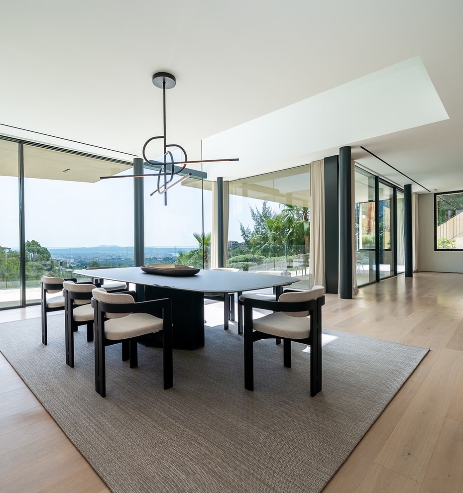 Dining room with an oval table and six chairs on a textured rug, floor-to-ceiling glass walls, and oak wood flooring.