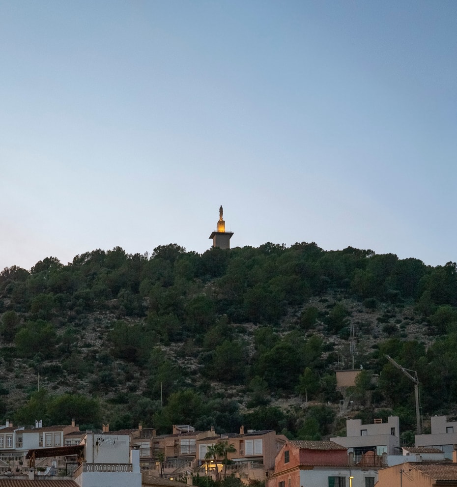 A distant view of a hillside topped with a tall landmark tower against a clear blue evening sky, seen above local rooftops.