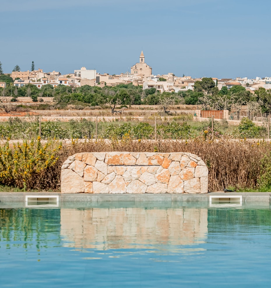A rectangular swimming pool featuring a natural stone waterfall wall with the village of Santanyí in the background.