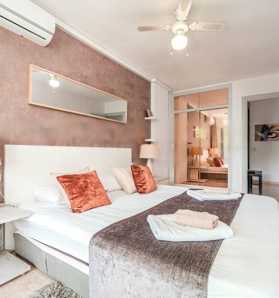 A bedroom featuring a textured dusty rose accent wall, a white bed with orange decorative pillows, a ceiling fan, and a view into a secondary room through an open doorway.