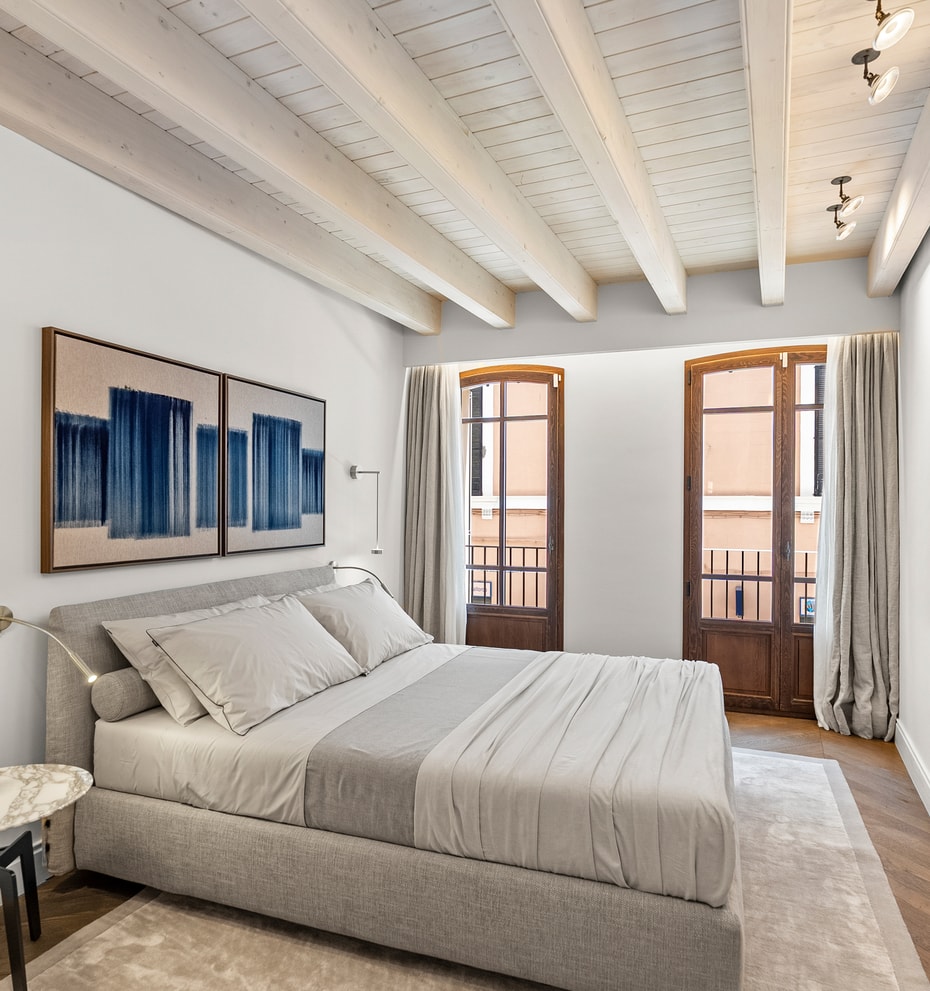 A bedroom featuring exposed timber ceiling beams, herringbone oak floors, and two arched glass doors opening to balconies.