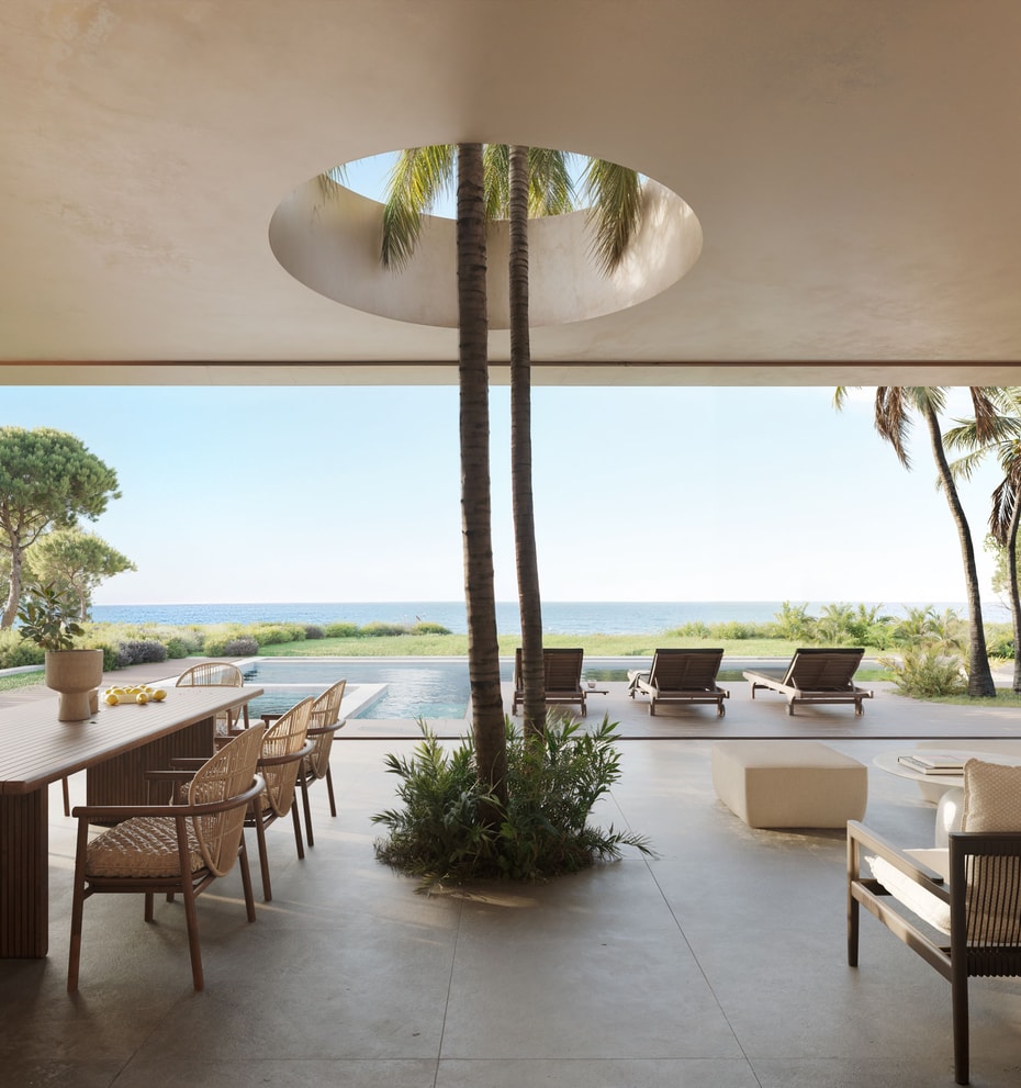 A wide outdoor covered terrace featuring a dining area, lounge seating, and an open circular skylight with trees, facing the sea.